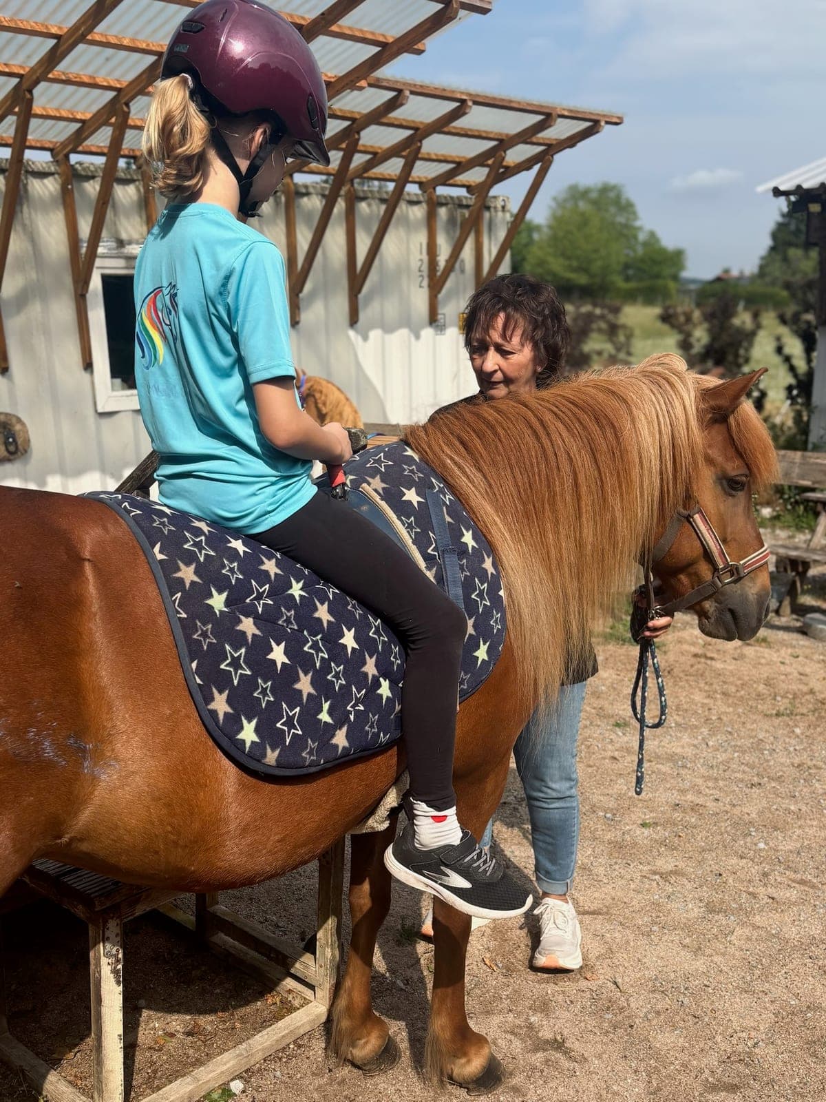 Kind sitzt auf einem Pony, das am Hof von einer Mitarbeiterin gehalten wird