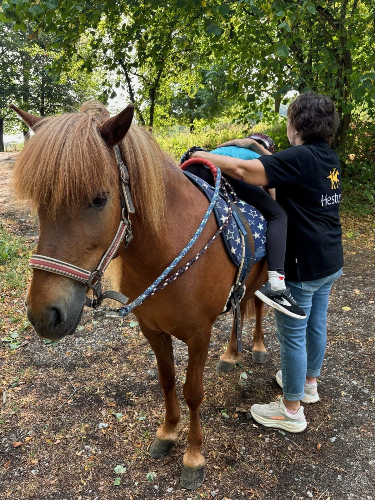 Mitarbeiterin sichert von hinten ein Kind auf einem Fuchspony auf einem schattigen Waldweg