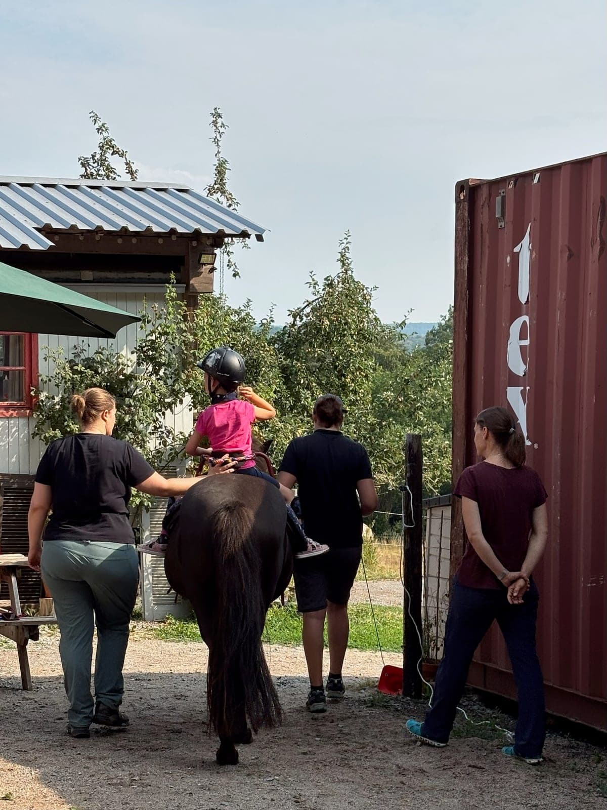 Ein Kind mit Reithelm reitet auf einem Pony, zwei Begleiterinnen führen es über das Hofgelände